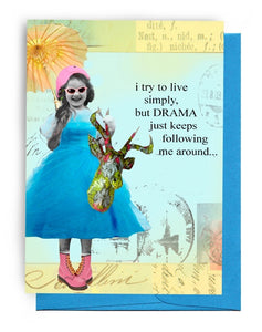 5" BY 7" GREETING CARD |  BLUE ENVELOPE. A GIRL HAS ON A BLUE, TAFFETA DRESS WEARING PINK, HIGH-TOP, LACE-UP BOOTS | PARASOL RIGHT HAND |  DECOPAGED DEER HEAD WITH ANTLERS IN HER LEFT | PINK HAT WITH PINK SUNGLASSES. BACKGROUND IS A LIGHT / BLUE TRAVEL STAMPS ALL OVER IT. WORDS OUTSIDE: ‘I TRY TO LIVE SIMPLY, BUT DRAMA JUST KEEPS FOLLOWING ME AROUND…’; INSIDE IS BLANK.