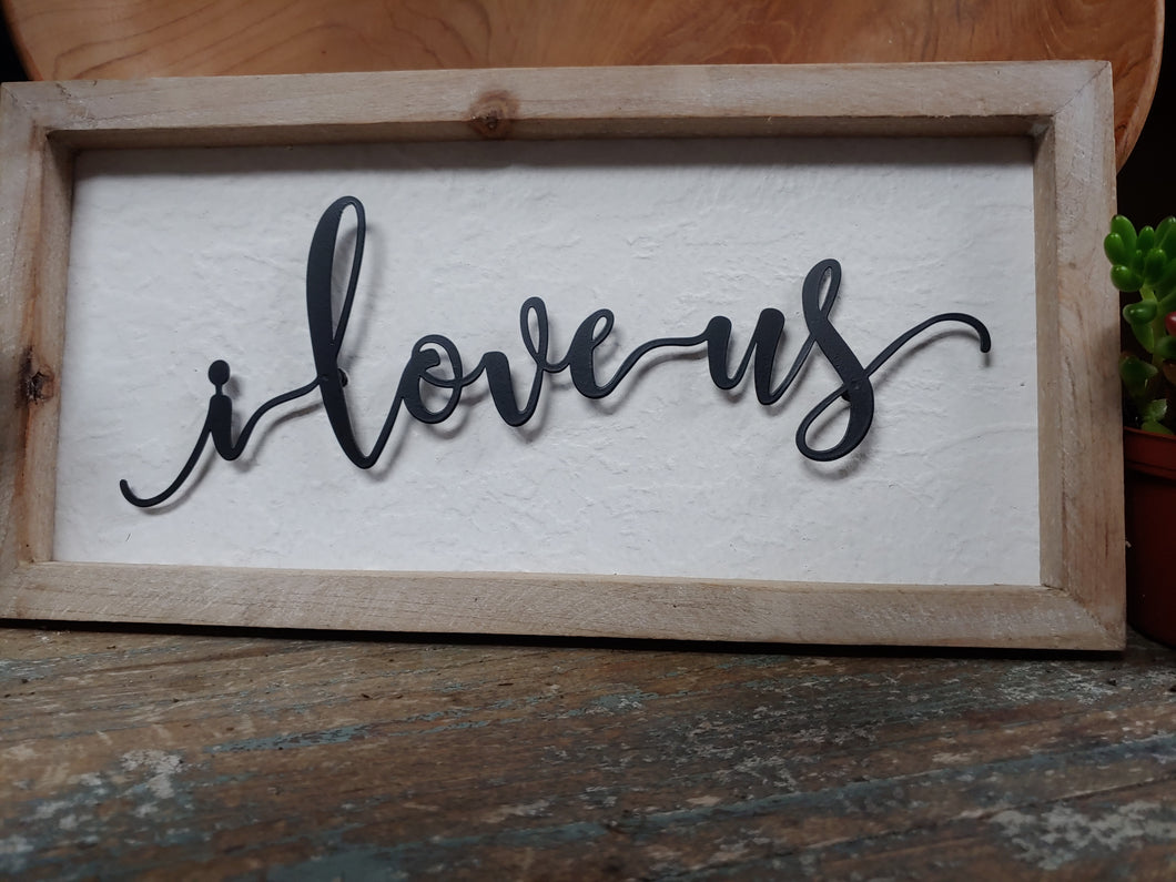 WOODEN [NATURAL FINISH] SHADOW BOX SIGN. THE BLACK IRON SCRIPT “I LOVE US” IS SUSPENDED ON A WHITE BACKGROUND—A FUN SIGN FOR MANY OF US.