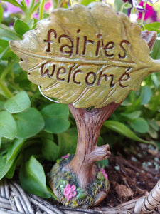 FAIRY GARDEN – RESIN – ‘FAIRIES WELCOME’ LEAF SIGN. 3” BY 3”, YELLOW-GREEN LEAF ON TOP OF TREE TRUNK THAT IS SURROUNDED BY PINK FLOWERS AND MOSS AT ITS BASE.