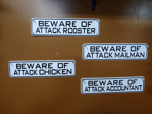4 BEWARE OF ATTACK SIGNS ON A BROWN WALL. THEY ARE BLACK WORDS ON WHITE METAL. 12 INCHES LONG BY 3” HIGH. SAYINGS ARE ‘BEWARE OF ATTACK ROOSTER, CHICKEN, MAILMAN, AND ACCOUNTANT.