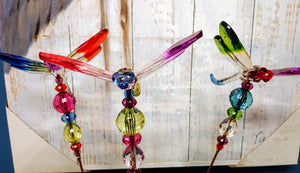 IMAGE OF 3 DRAGONFLIES AGAINST A LIGHT-WOOD WALL, ALL WITH CLEAR BODIES. EACH DRAGONFLY HAS DIFFERENT COLORED WINGS, TAILS, AND HEADS. BEADS ON THE STAKES ARE RED, BLUE, GREEN, CLEAR, AND GOLD.  RedShedGarden redshedgarden RedShedredshed Baraboo Gifts Mother’s Day Birthday Anniversary Garden Stakes Dragonflies Just Because