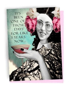 A GREETING CARD, PINK ENVELOPE. WOMAN WITH DARK SHOULDER-LENGTH HAIR, PINK FLOWER, AND WHITE FLOWER ON BOTH SIDES OF HEAD, WEARING, WHITE-FRAMED GLASSES. WEARING TREE-STAMPED WHITE DRESS. BLACK NAIL POLISHED HAND COMES FROM BOTTOM HOLDING A MARTINI. TOP LEFT HAS BLUE BACKGROUND; WORDS OUTSIDE -- IT’S ‘ONE OF THOSE DAYS’ FOR LIKE 3 YEARS NOW…INSIDE I’M BEGINNING TO THINK IT MAY JUST BE ME! 