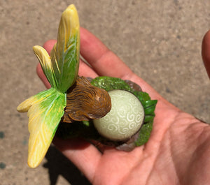 FAIRY GARDEN – RESIN – ‘MAGICIAN’ | TOP VIEW OF FAIRY GIRL/LIGHT GREEN WINGS WITH YELLOW EDGES/LONG BROWN HAIR | LEANING OVER A ‘GLOW-IN-THE-DARK’ CRYSTAL BALL – CAN SEE ‘MAGIC’ SWIRLS/IN METAL-BANDED STAND/ON ROCKS-GREEN MOSS-GREEN LEAVES | SITTING ON A PERSON’S HAND.