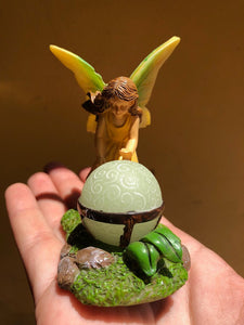 FAIRY GARDEN – RESIN – ‘MAGICIAN’ | FRONT VIEW OF FAIRY GIRL/LIGHT GREEN WINGS WITH YELLOW EDGES/LONG BROWN HAIR/SLEEVELESS, YELLOW DRESS | LEANING OVER A ‘GLOW-IN-THE-DARK’ CRYSTAL BALL/IN METAL-BANDED STAND – CAN SEE ‘MAGIC’ SWIRLS/ON ROCKS-GREEN MOSS-GREEN LEAVES | SITTING ON A PERSON’S HAND.