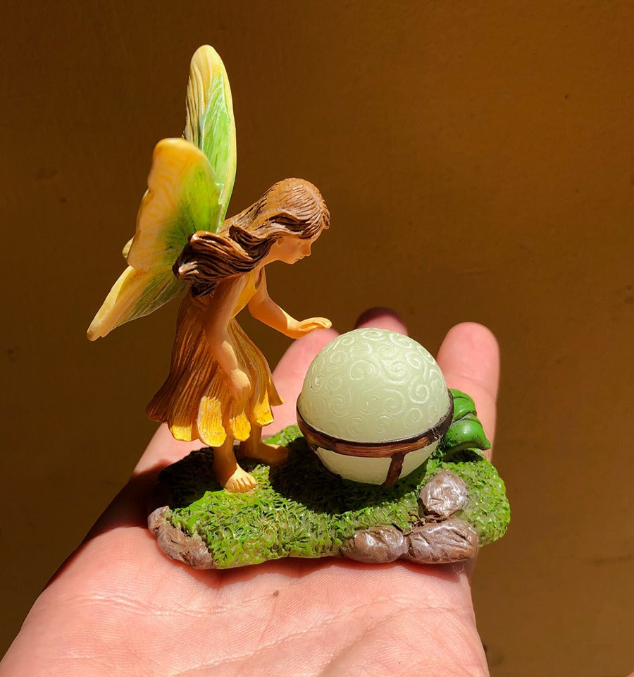 FAIRY GARDEN – RESIN – ‘MAGICIAN’ | LEFT SIDE OF FAIRY GIRL/LIGHT GREEN WINGS WITH YELLOW EDGES/LONG BROWN HAIR/SLEEVELESS, YELLOW DRESS | LEANING OVER A ‘GLOW-IN-THE-DARK’ CRYSTAL BALL/IN METAL-BANDED STAND/ON ROCKS-GREEN MOSS-GREEN LEAVES | SITTING ON A PERSON’S HAND.