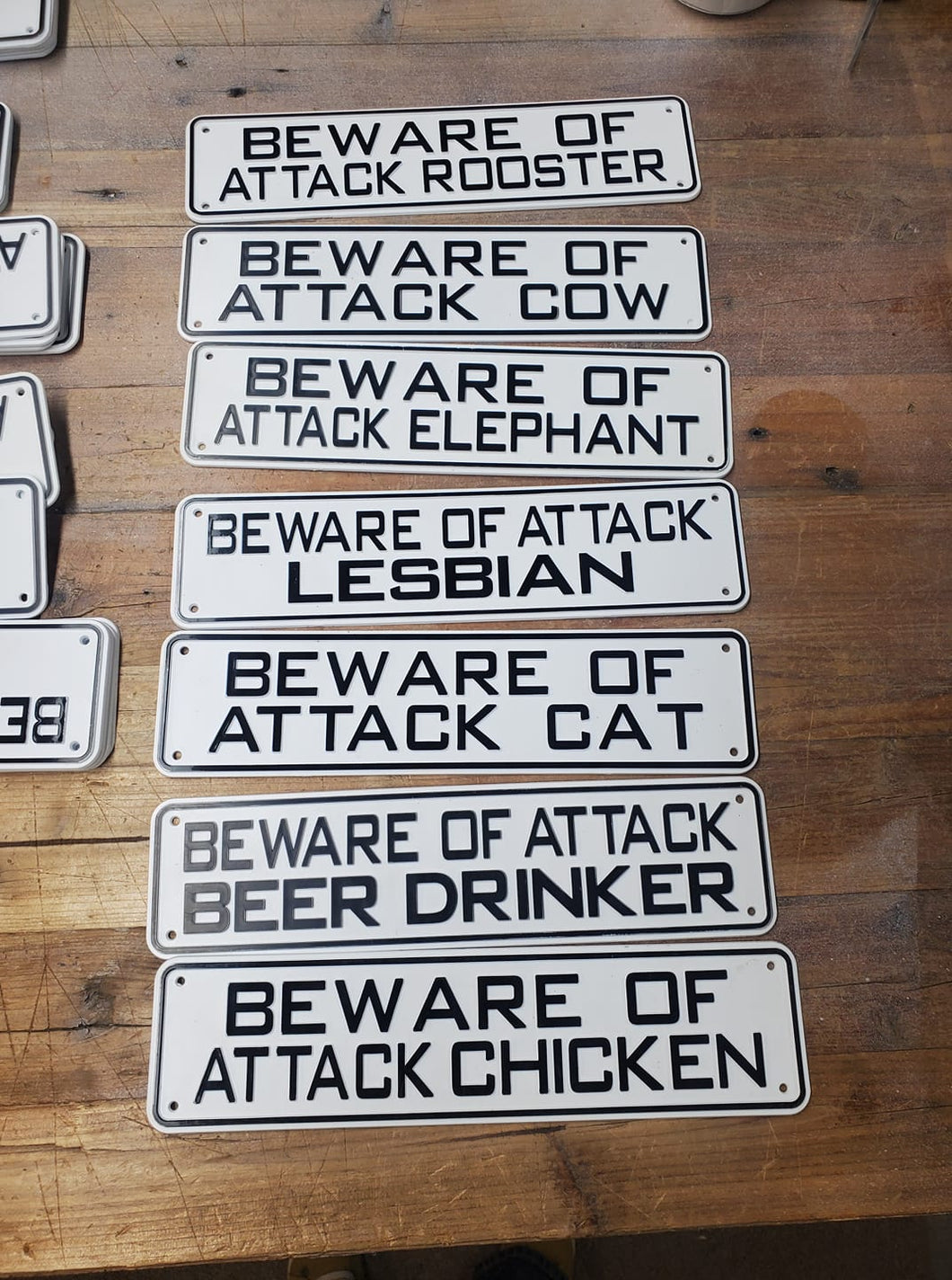 7 BEWARE OF ATTACK SIGNS ON A WOODEN TABLE. THEY ARE BLACK WORDS ON 12 INCHES LONG BY 3” HIGH WHITE METAL. SOME SAYINGS ARE ‘BEWARE OF ATTACK ROOSTER, COW, ELEPHANT, CAT, CHICKEN, MOSQUITO, MUSICIAN, GARDNER,NO PEEING,KEEP OUT