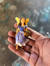 Load image into Gallery viewer, FAIRY GARDEN – RESIN – NEW! ‘BLOSSOM FAIRY’ | MG322 | 3.5” T | YOUNG LADY FAIRY | LIGHT GOLDEN WINGS/LONG BROWN HAIR/SHORT-SLEEVED, FLOOR-LENGTH DRESS – DARK PURPLE FADING TO WHITE ON BOTTOM, WHITE BELTED WAISTLINE | CARRYING BROWN, WOVEN BASKET ON LEFT ARM/WHITE, PURPLE, YELLOW, AND PINK FLOWERS/BLACK STAKE ON BOTTOM TO HELP STAND. LAYING ON A PERSON’S FINGERS TO SHOW SIZE.