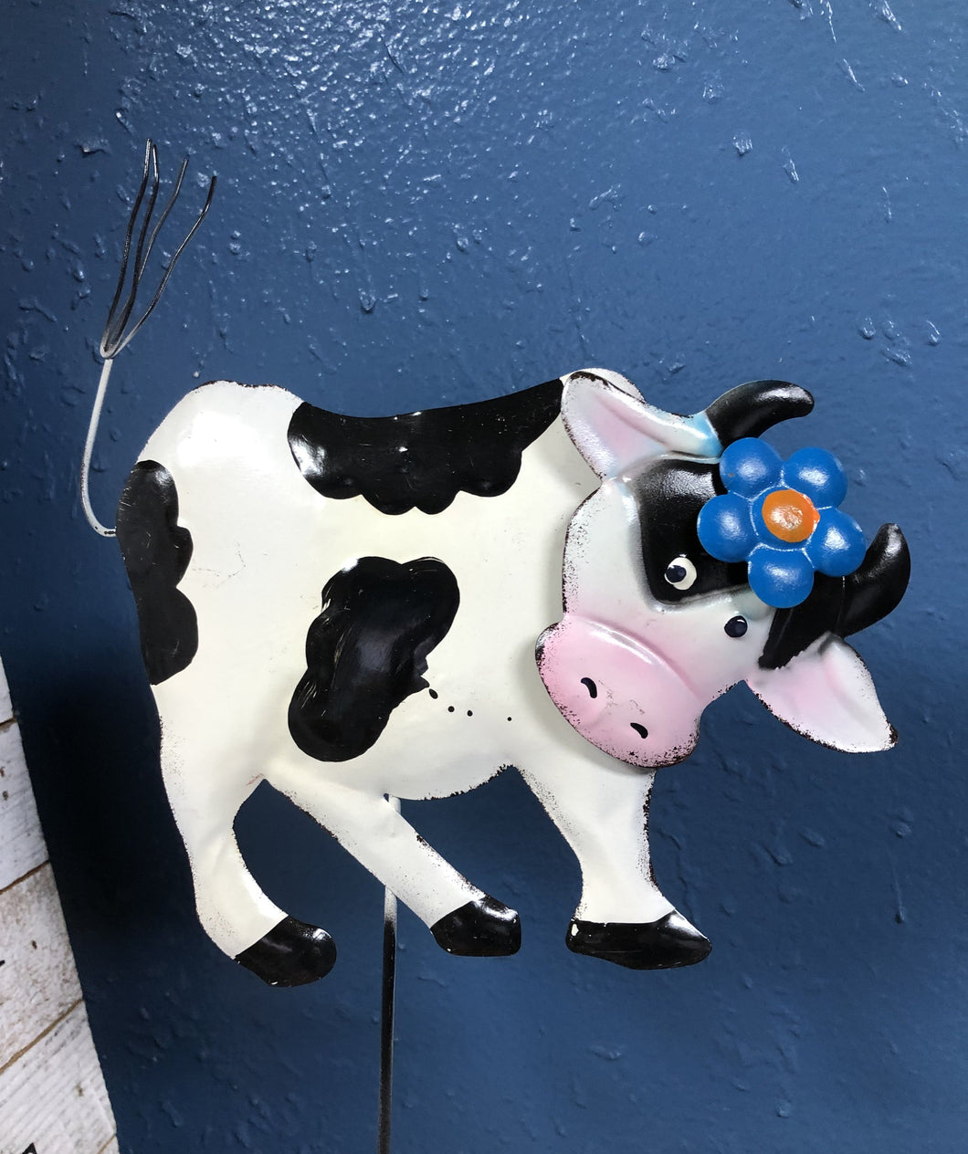 METAL COW 6 INCHES TALL BY 8 INCHES WIDE ON A METAL STAKE. COLORS ARE BLACK AND WHITE COW WITH BLACK HORNS AND A BLUE FLOWER ON ITS HEAD, AGAINST A BLUE WALL.