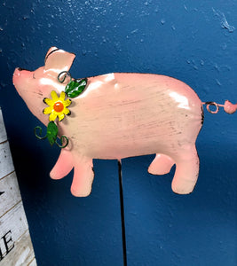 METAL PIG 6 INCHES TALL BY 8 INCHES WIDE ON A METAL STAKE. COLORS IS ALL PINK WITH A CURLY TAIL WEARING A YELLOW FLOWER WITH GREEN VINES AROUND ITS NECK, AGAINST A BLUE WALL.