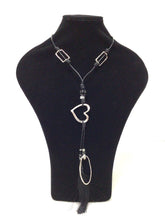 Load image into Gallery viewer, A modern, stylish necklace.  Pendant features an open heart, an elongated oval and a black silklike tassel. There are two 1.25&quot;L open rectangles that adorn the black leather-like cord necklace. The cords also have lovely ceramic, and silver spacers. Can slip easily over the head but also has a lobster claw clasp and an additional 2&quot; of chain for added length. 