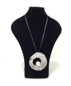 3 black slender cord strands hold a substantial silver tone circle pendant that has an open center. On the bottom of the center hole sits a large white faux pearl approximately nickel sized. The surface of the metal is mottled and there is a clasp on the cord. 