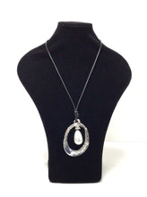 Load image into Gallery viewer, Pendant necklace has an elongated oval in silver-tone, mottled metal. The oval pendant  is accentuated with a large single teardrop-shaped faux pearl that hands in the oval. The pendant is hung on a simple black braided cord that does have a lobster claw clasp that can add an additional 2&quot; in length however it can easily slip over the head.