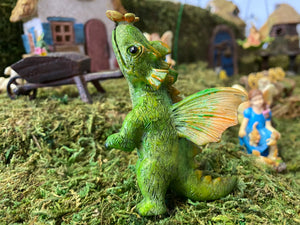 FAIRY GARDEN – RESIN – ‘LITTLE DRAGON’ FAIRY | MG305 | 3” T BY 2” W BY 2” D | 1” STAKE AT BOTTOM | GREEN SCALES/YELLOW-ORANGE WINGS/YELLOW-GREEN RIDGE-LINE SCALES | ON MOSS | ON BACK LEGS WITH YELLOW BUTTERFLY ON ITS NOSE | GREY WHEELBARROW IN BACKGROUND.