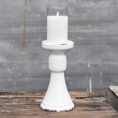 A 9 INCH TALL WHITE, CERAMIC, MINIMALISTIC-LOOKING PILLAR CANDLE HOLDER. IT IS 4.5 INCHES DEEP AND HAS A WHITE CANDLE ON TOP OF IT. SHOWS SLIGHT IMPERFECTIONS DUE TO BEING HAND-GLAZED. IT SITS ON A WOODEN PLATFORM WITH A BLURRED GREY WALL BEHIND IT.