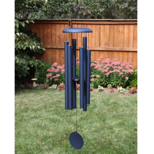 Load image into Gallery viewer, AN IODIZED, DARK BLUE, ALUMINUM WINDCHIME/WINDCATCHER. IT IS HANGING OVER A GRASS YARD. IT HAS 6 ALUMINUM POWDERCOATED TUBES, LARGE TOPPER, AND WINDCATCHER DOWN THE MIDDLE. BEHIND ARE TREES, A LARGE MUSHROOM STATUE UNDER A TREE, AND A FLOWER GARDEN ALONG A WOODEN PRIVACY FENCE.