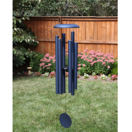 AN IODIZED, DARK BLUE, ALUMINUM WINDCHIME/WINDCATCHER. IT IS HANGING OVER A GRASS YARD. IT HAS 6 ALUMINUM POWDERCOATED TUBES, LARGE TOPPER, AND WINDCATCHER DOWN THE MIDDLE. BEHIND ARE TREES, A LARGE MUSHROOM STATUE UNDER A TREE, AND A FLOWER GARDEN ALONG A WOODEN PRIVACY FENCE.