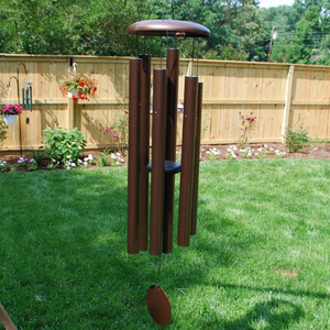 AN IODIZED, BRONZE, ALUMINUM WINDCHIME/WINDCATCHER. IT IS HANGING FROM A PLANT HANGER OVER A GRASS YARD. IT HAS 6 ALUMINUM POWDERCOATED TUBES, LARGE TOPPER, AND WINDCATCHER DOWN THE MIDDLE. BEHIND ARE TREES, POWERLINES, A SECOND WINDCHIME HANGING WITH A PLANT FROM A PLANT HANGER, AND A FLOWER GARDEN ALONG A WOODEN PRIVACY FENCE.