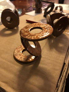 4 METAL AIR PLANT STANDS. 3 LARGE WASHERS WELDED TOGETHER TO FORM A ‘Z.’ SITTING ON A BOX. BEHIND ARE 3 MORE ‘WELDED WASHERS’ PLANT STANDS FORMED INTO CIRCLES. ALL ARE PAINTED METALLIC COPPER. 