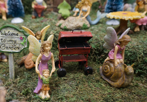 FAIRY IN A PINK DRESS KNEELING ON A BROWN SNAIL|3 INCHES TALL. THEY ARE IN THE FAIRY GARDEN WITH FAIRY MOM IN A PURPLE DRESS WALKING FAIRY BOY IN A GREEN SHIRT AND BROWN SHORTS|RED GRILL|SIGN ‘WELCOME TO MY GARDEN’.
