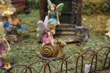 Load image into Gallery viewer, FAIRY IN A PINK DRESS KNEELING ON A BROWN SNAIL|3 INCHES TALL|HAS IRON FENCE IN FRONT. THEY ARE IN THE FAIRY GARDEN WITH FAIRY IN BLUE DRESS SITTING ON WATER FOUNTAIN STONES.