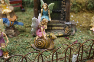 FAIRY IN A PINK DRESS KNEELING ON A BROWN SNAIL|3 INCHES TALL|HAS IRON FENCE IN FRONT. THEY ARE IN THE FAIRY GARDEN WITH FAIRY IN BLUE DRESS SITTING ON WATER FOUNTAIN STONES.