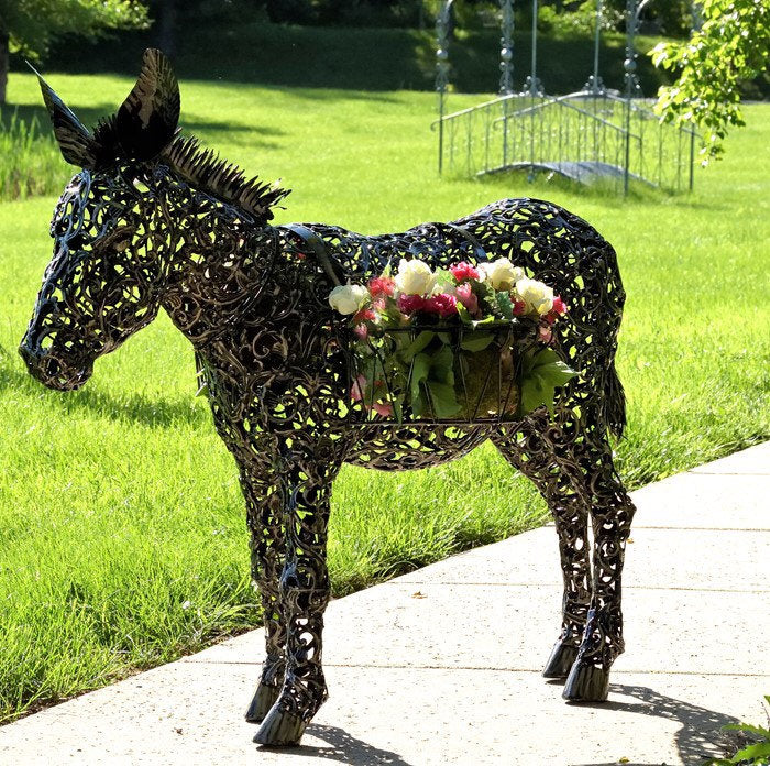 AN ANTIQUE BRONZE-FINISHED, 3 FOOT FILIGREE METAL DONKEY WITH BASKETS. DONKEY IS ON SIDEWALK NEXT TO A LAWN. THERE IS A METAL WALK BRIDGE IN THE BACKGROUND.
