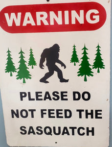 White metal sign that reads Warning!  Please do not feed the sasquatch with a picture of big foot and 6 evergreen pine trees