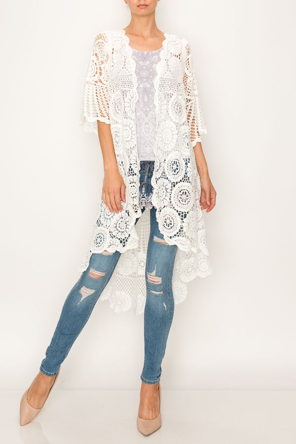 WOMAN WEARING BRIGHT WHITE, CROCHETED DUSTER JACKET | PAIRED TO A WHITE, SHORT-SLEEVED BLOUSE WITH GREY PATTERN | WEARING SKINNY LEG JEANS WITH FASHIONABLE TEARS IN THE LEGS AND LIGHT TAN HIGH HEELS.